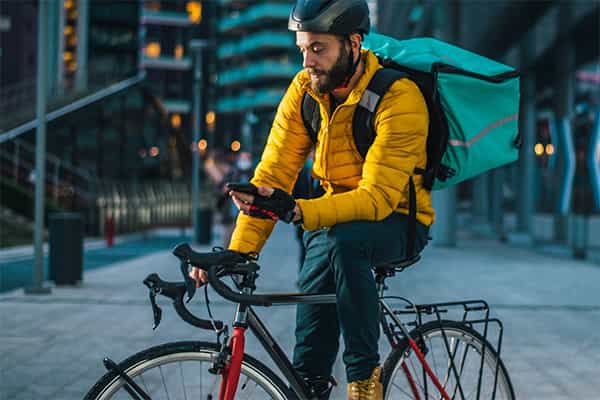 Livreur à vélo portant une veste jaune et un sac à dos sarcelle regardant son téléphone dans une rue urbaine